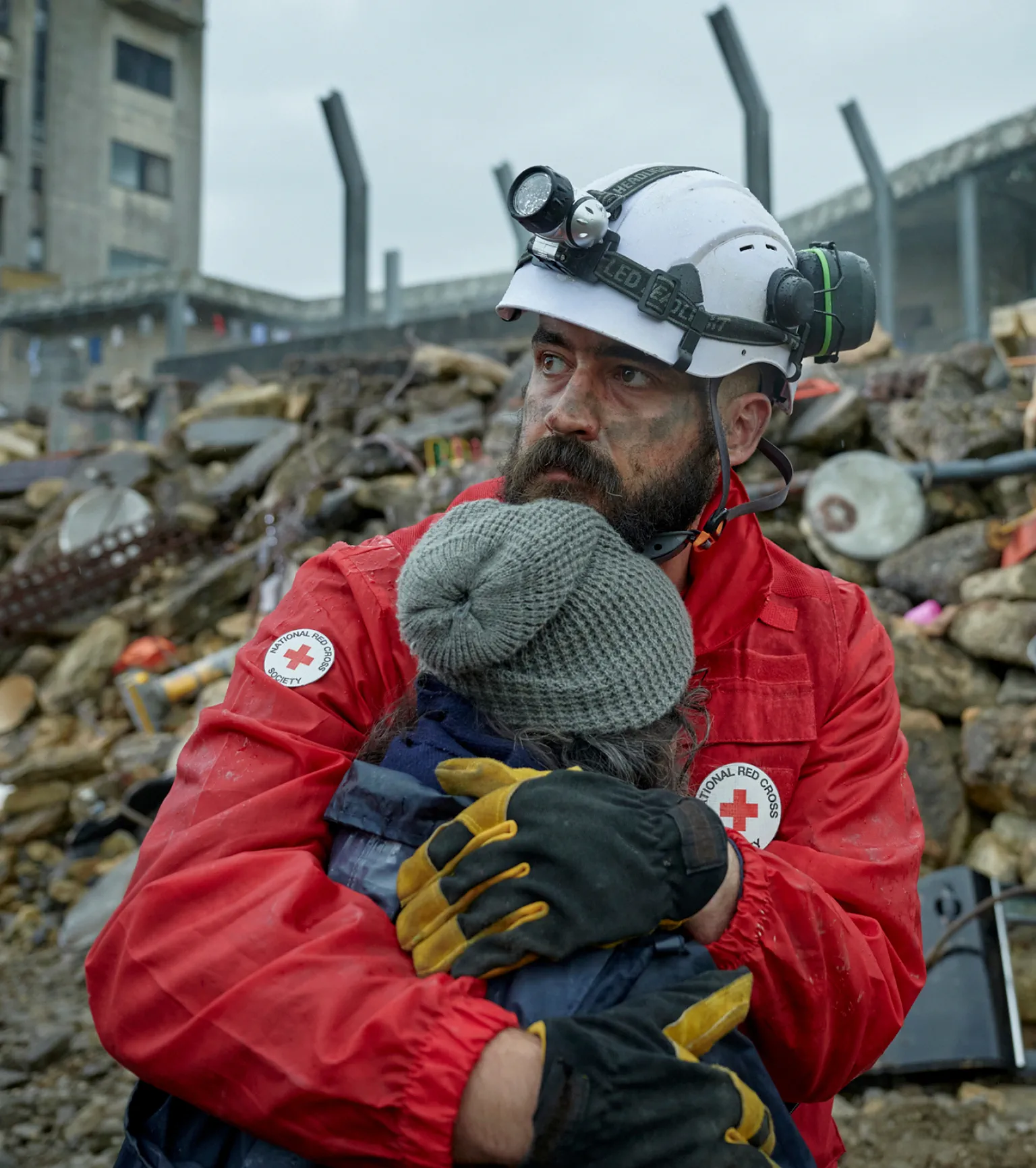 A Red Cross worker in red jacket and helmet hugs a person wearing a winter coat and hat, with rubble from a destroyed building in the background.