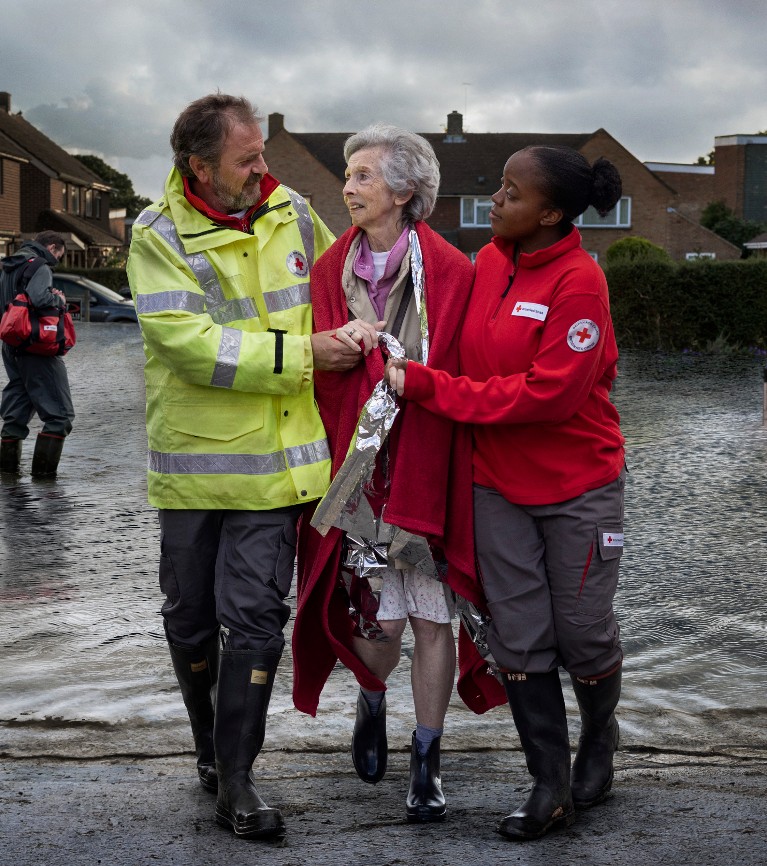 Two British Red Cross volunteers help a woman whose home has been flooded. She is wrapped in a blanket as they wade through flood water together