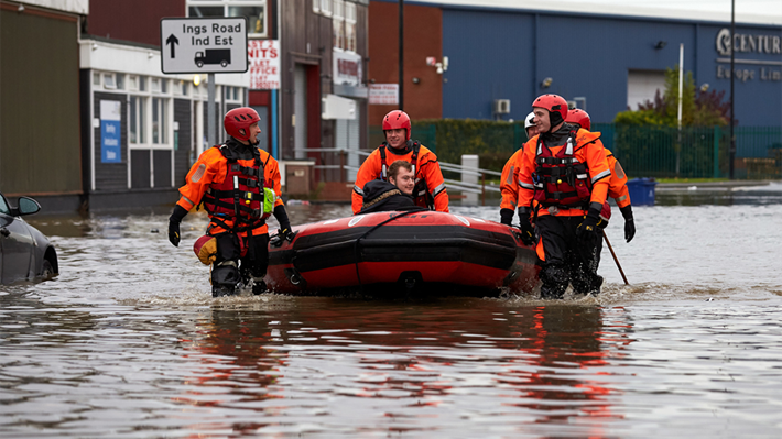 In a flooded street in the UK, Red Cross volunteers wearing protective clothing and boots help by pulling an inflatable raft with a man sitting in it through the water