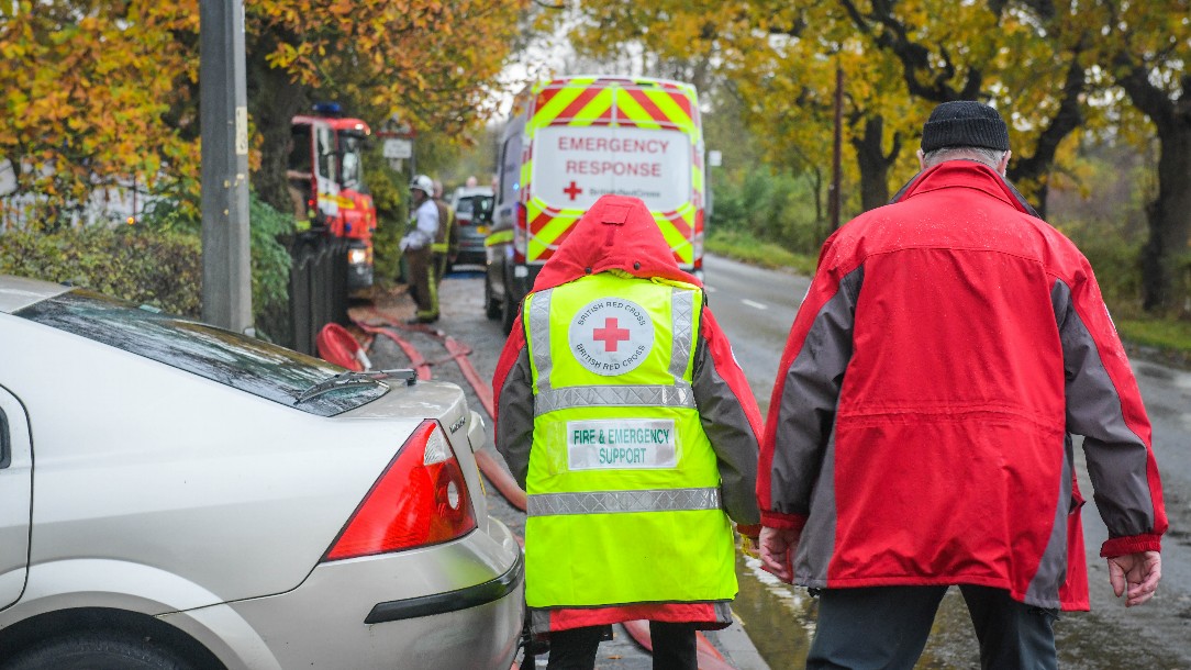 Two British Red Cross volunteers walk away from the camera, wearing their uniforms. In the background, people are carrying out flood rescue work.