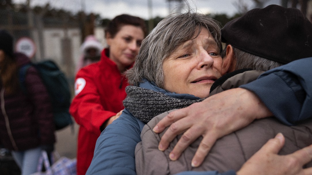 A woman hugs a relative who has their back to the camera. In the background, a Red Cross volunteer puts a comforting hand on her shoulder
