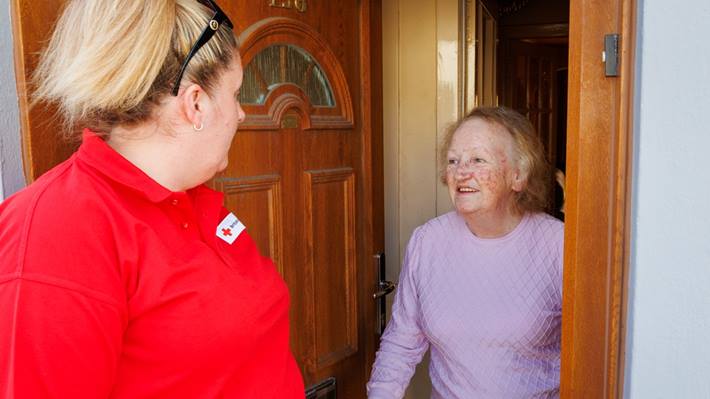 A photo of a Red Cross volunteer, in a red shirt, greeting an older lady at her front door.