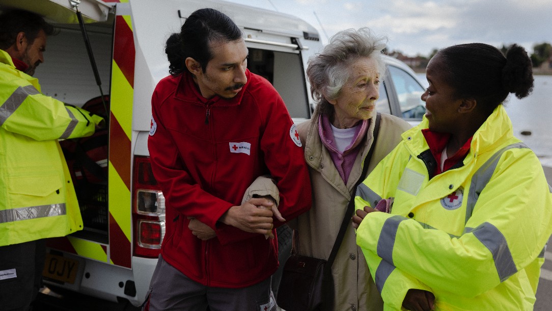 A woman is helped by two Red Cross volunteers, a man in a red Red Cross fleece and a woman in a hi-vis jacket. Their arms are linked and there is a Red Cross emergency van, along with a male volunteer, behind them