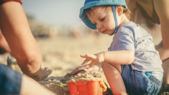 A young child plays with a bucket and spade at the beach with their family.