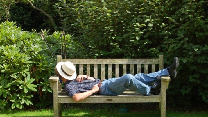 Someone asleep on a garden bench with a sun hat on their face.