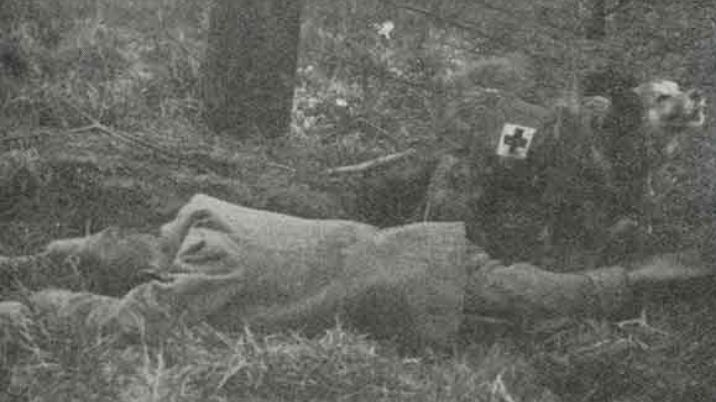 A black and white photograph showing an injured man and a dog wearing a British Red Cross coat barking for help.