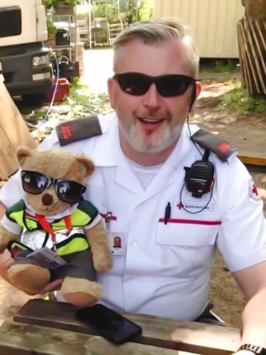 Volunteer Mike smiles at the camera wearing his Red Cross uniform and holding a teddy bear dressed in Red Cross colours