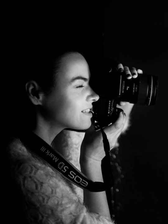 Black and white portrait of a woman (Olena) seen from the side, holding up a camera to her face to take a photograph