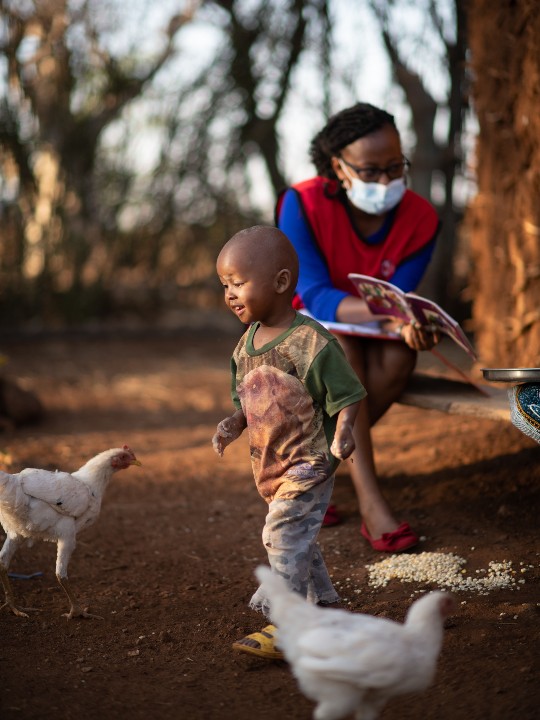 Member of the Kenyan Red Cross team points at book while child plays nearby.