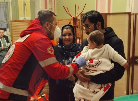 A Polish Red Cross volunteer speaks to a family with a baby who is wrapped in a Polish Red Cross blanket
