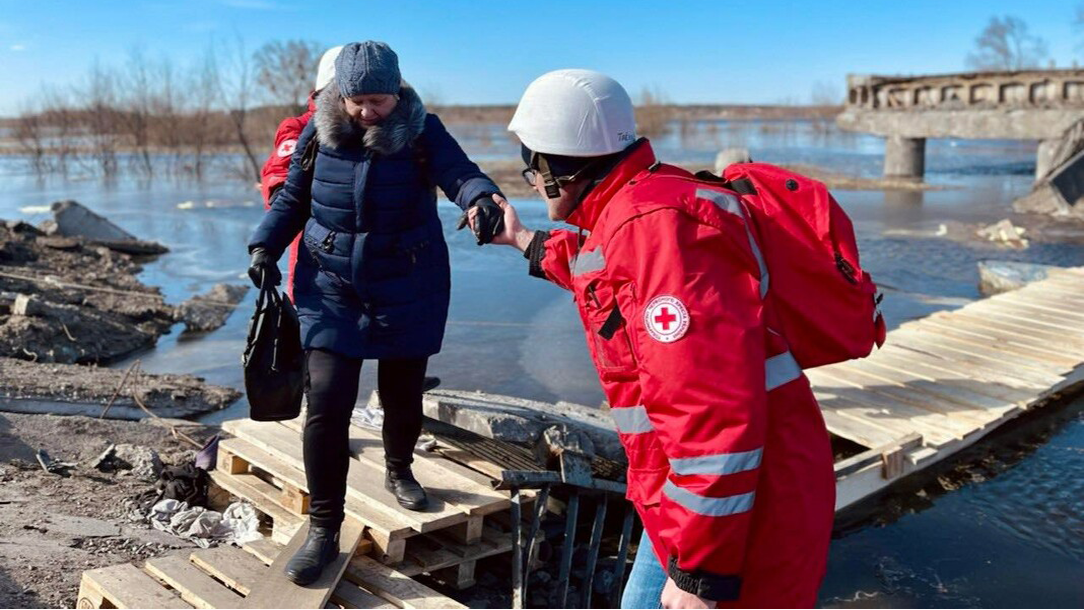 A Red Cross volunteer holds the hand of a woman crossing a makeshift wooden bridge while carrying a bag in Ukraine.