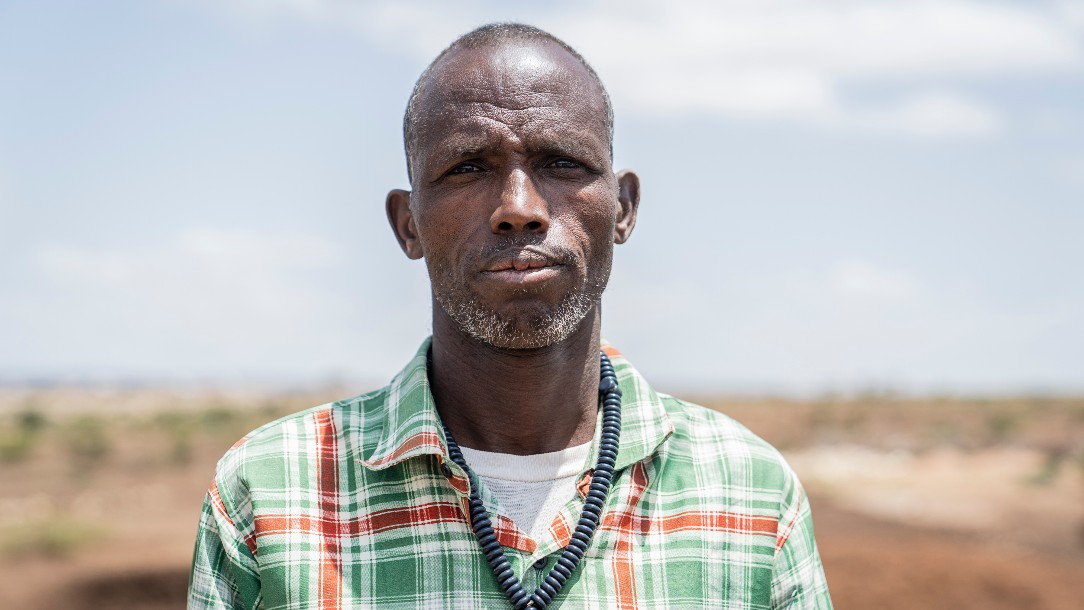 A man in a green and red checked shirt is pictured from the shoulders up, looking at the camera and standing in front of a dry landscape