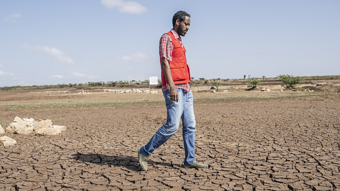 A man wearing a Red Cross vest walks across dry, cracked ground in Africa.