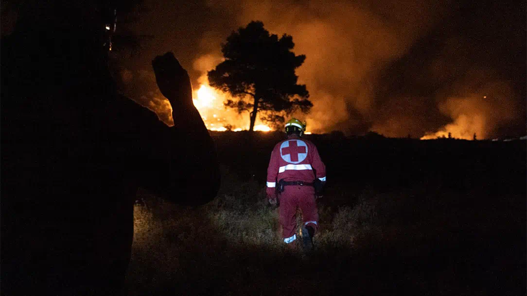 A person in a Red Cross uniform walks towards a wildfire at night. 