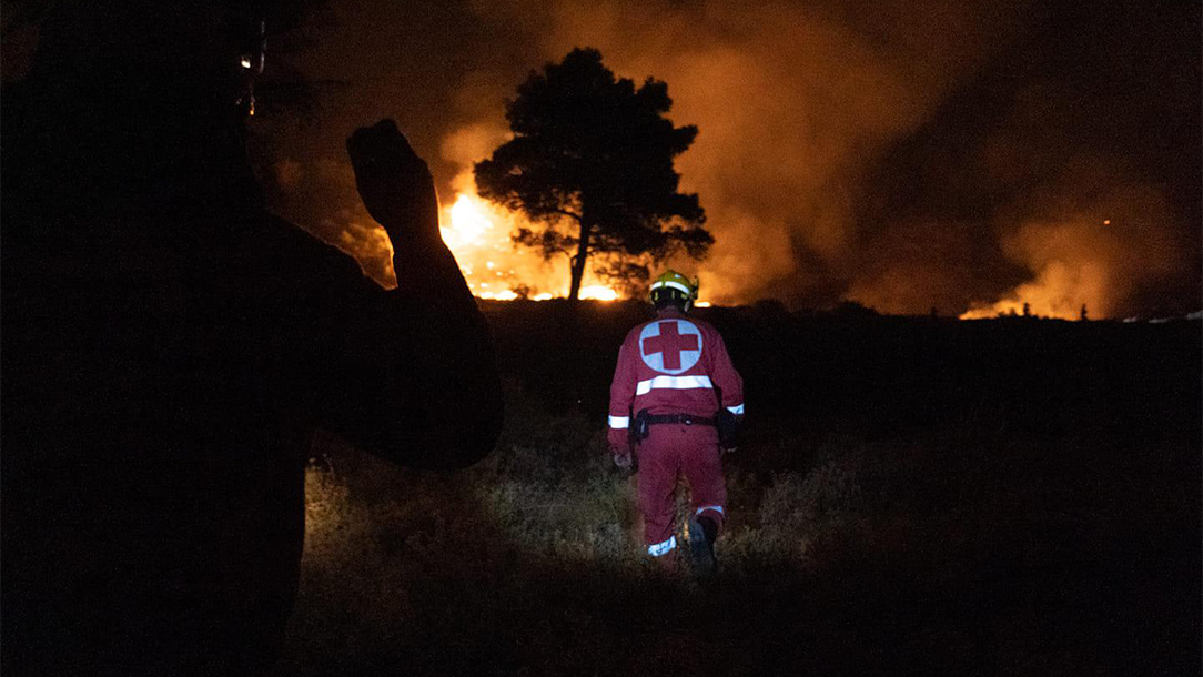 A person in a Red Cross uniform walks towards a wildfire at night.