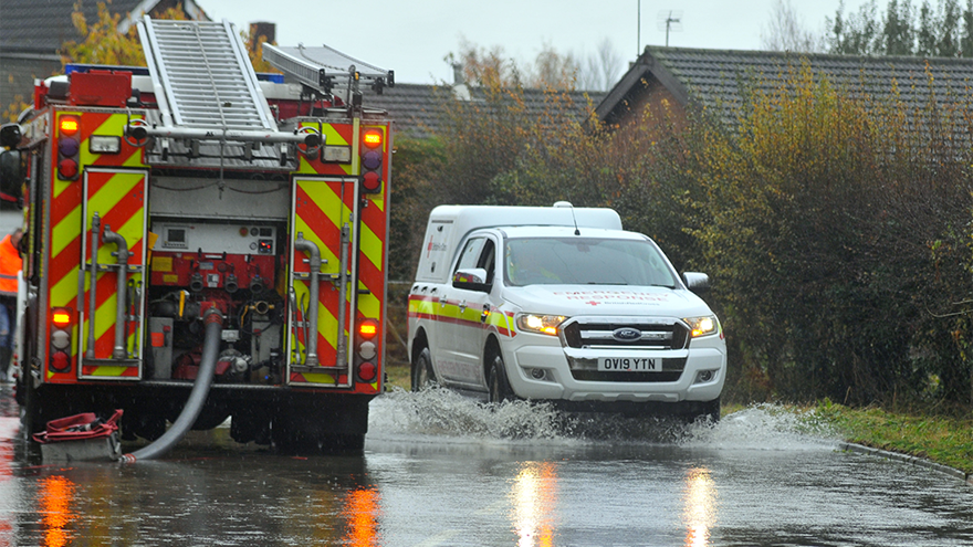 A fire engine and a British Red Cross emergency vehicle drive through a flooded road.