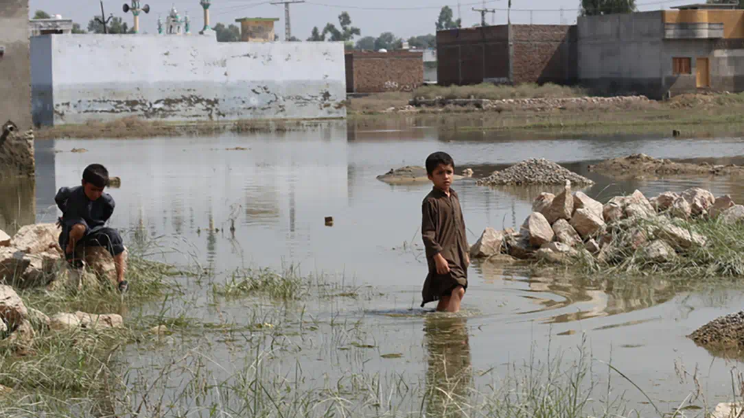 In Pakistan, two boys stand in flood water with rubble in the background.