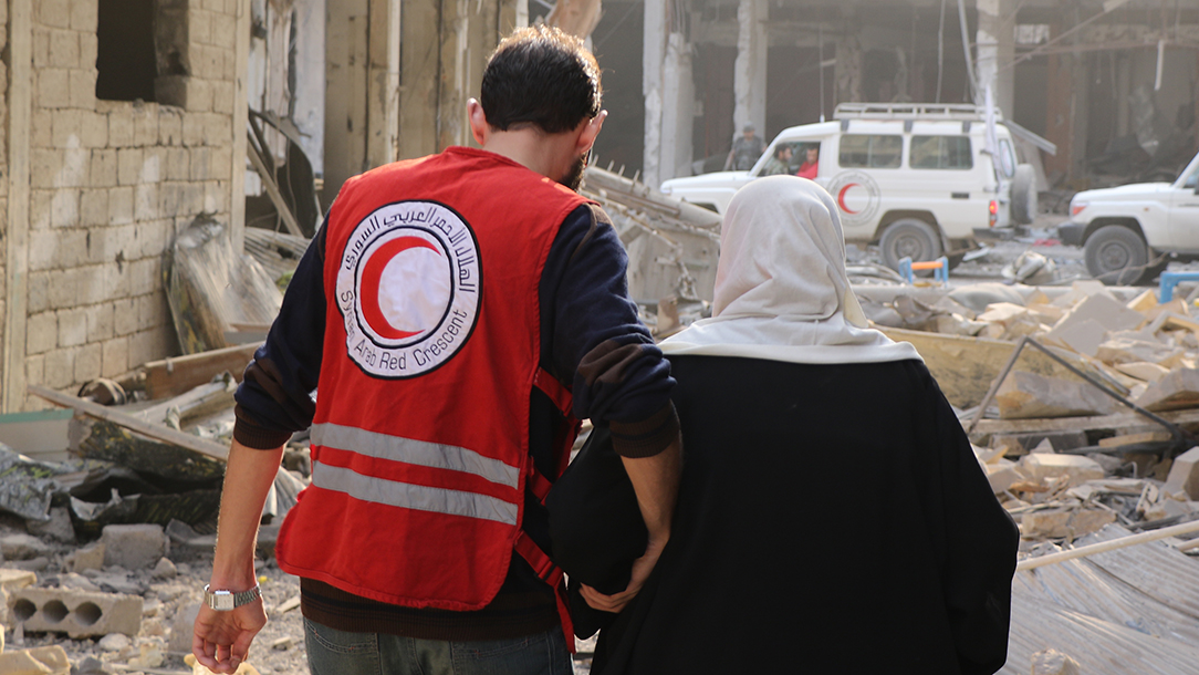 A volunteer from British Red Cross partners the Syrian Arab Red Crescent helps an older woman through a street full of rubble.