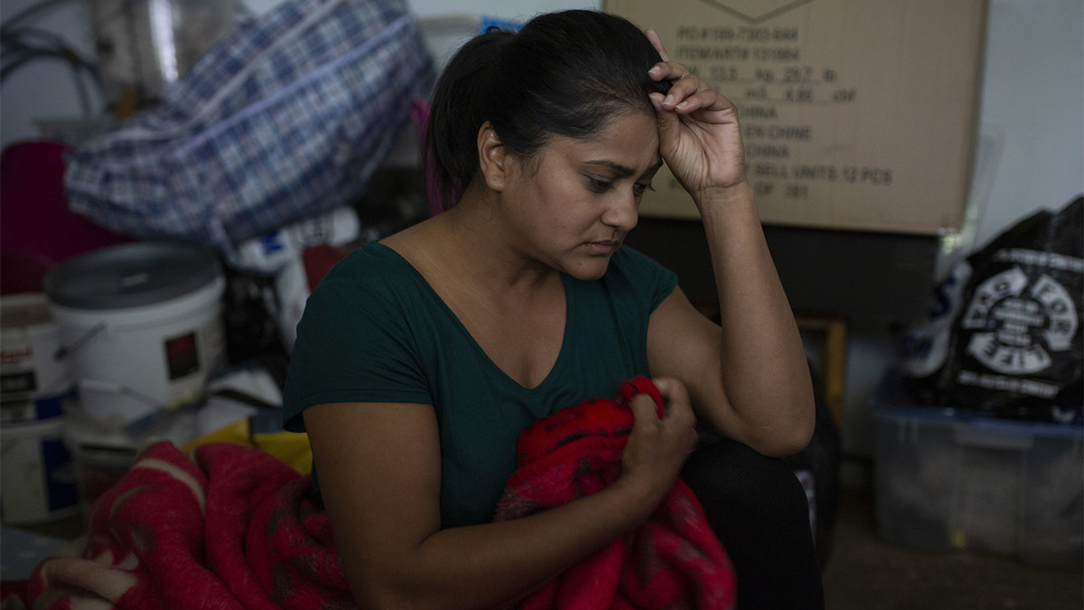 A woman sits in a small space surrounded by storied items with her hand held up to her head, looking sad. 