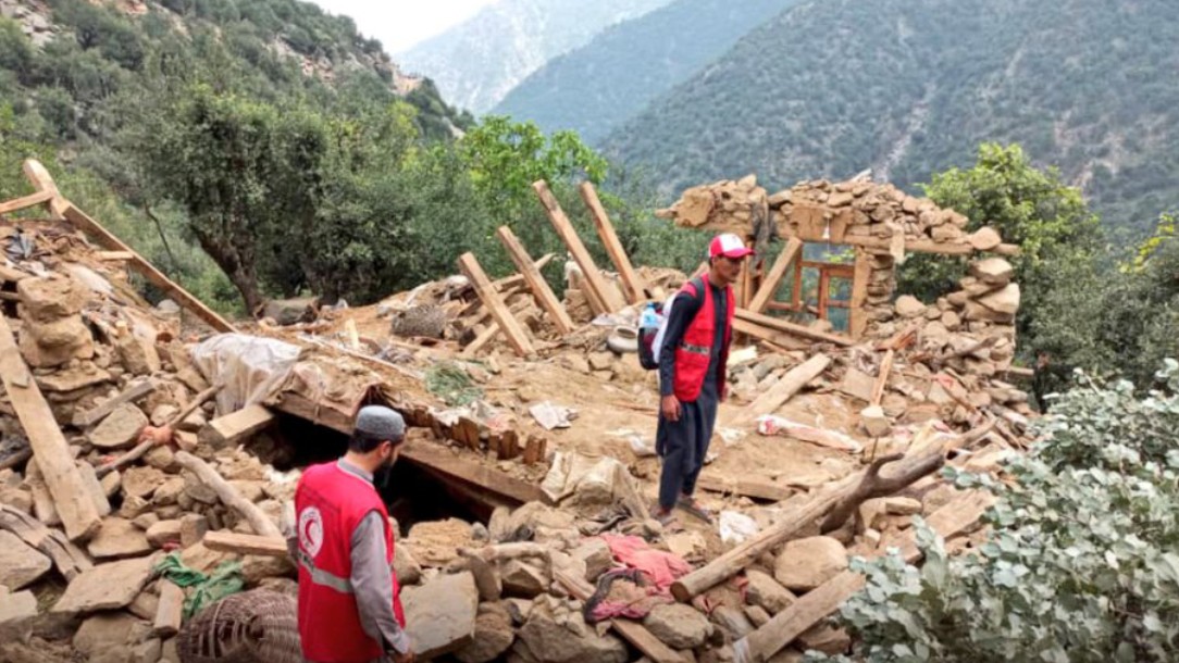 Two men wearing Red Cross uniforms stand in the rubble of a building destroyed by the 2025 earthquake in Afghanistan. There are mountains in the background.