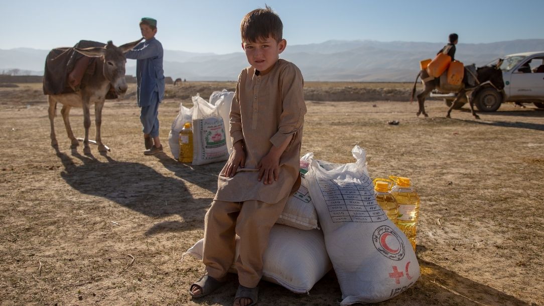 A small, sad looking boy sits on a bag of supplies in the middle of an arid landscape.