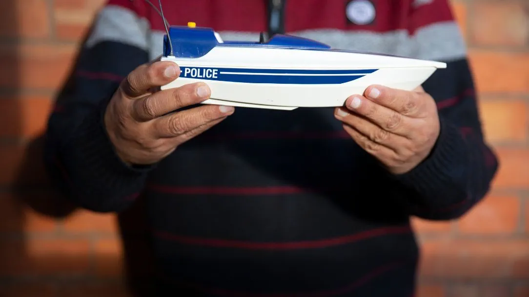 A man holds a small model police boat up to the camera.