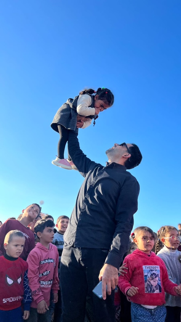 Amr, a PRCS media officer, pictured in a group of children while holding a young girl in the air playfully.