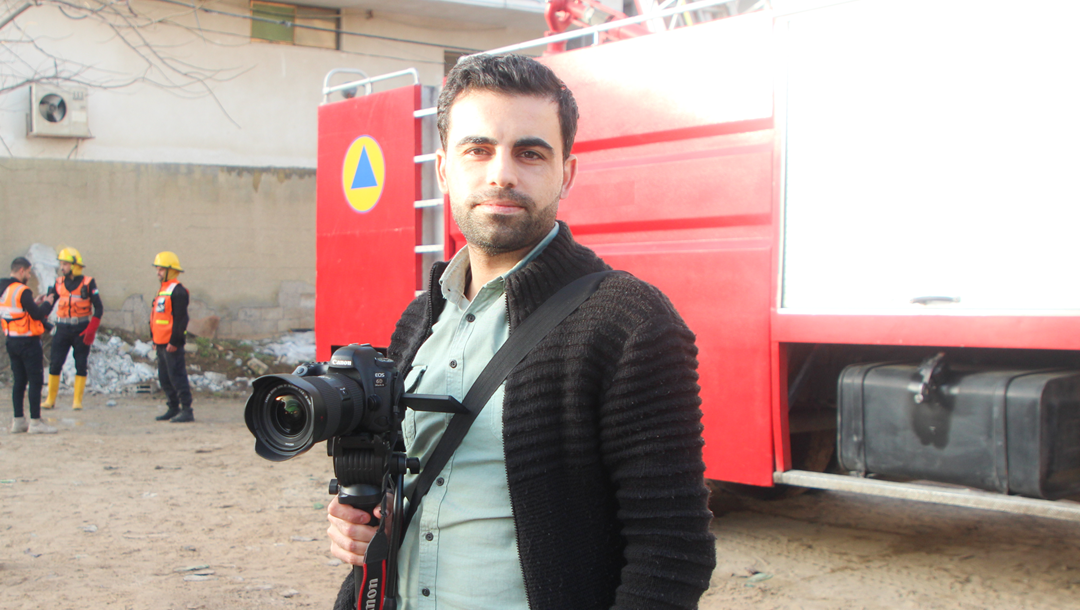A photo of Amr, a PRCS media officer who lives in Gaza. He is standing, holding his camera, in front of an emergency rescue vehicle.