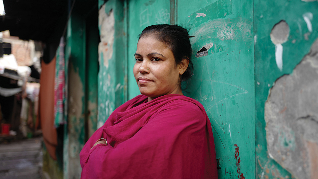 In Bangladesh, Josna stands against a wall smiling with her arms folded,