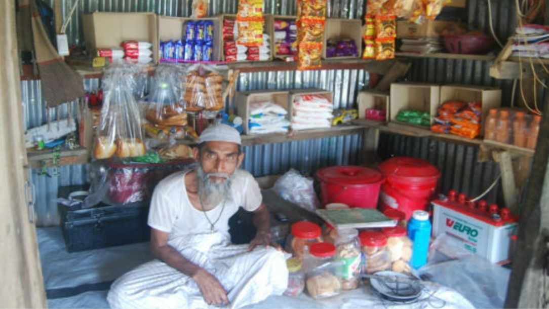 Man in Bangladesh sits on the floor of his shop, surrounded by goods.