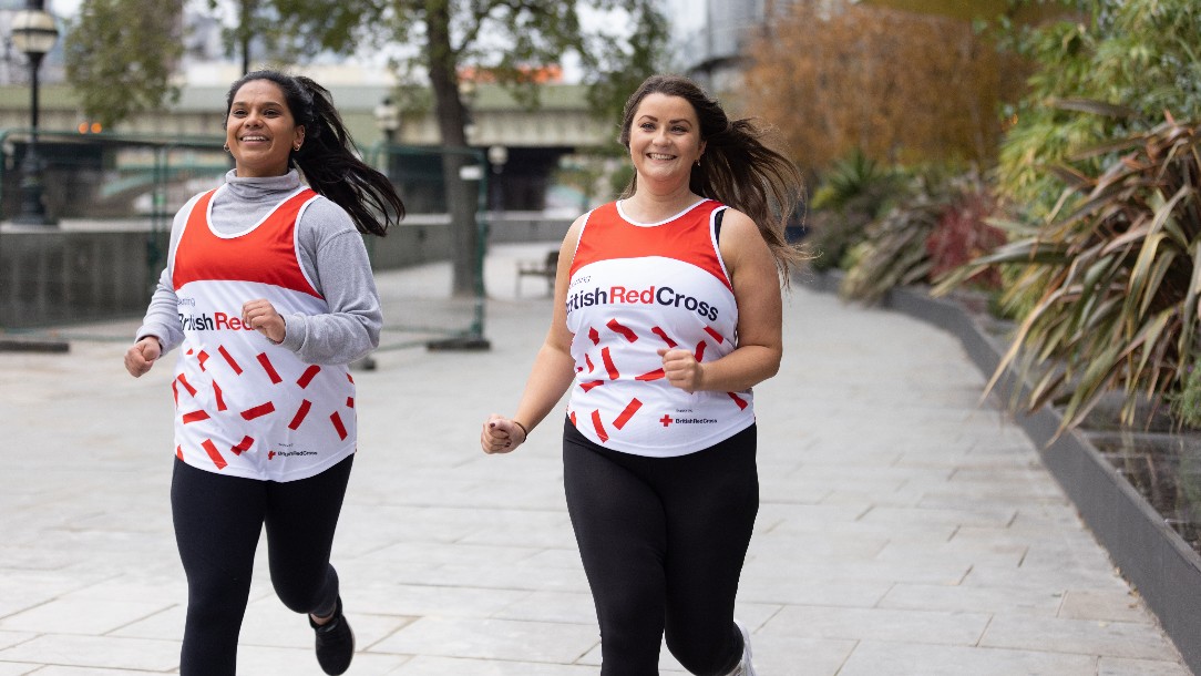 Two women wearing running gear including British Red Cross branded race vests, run along the pavement alongside the River Thames
