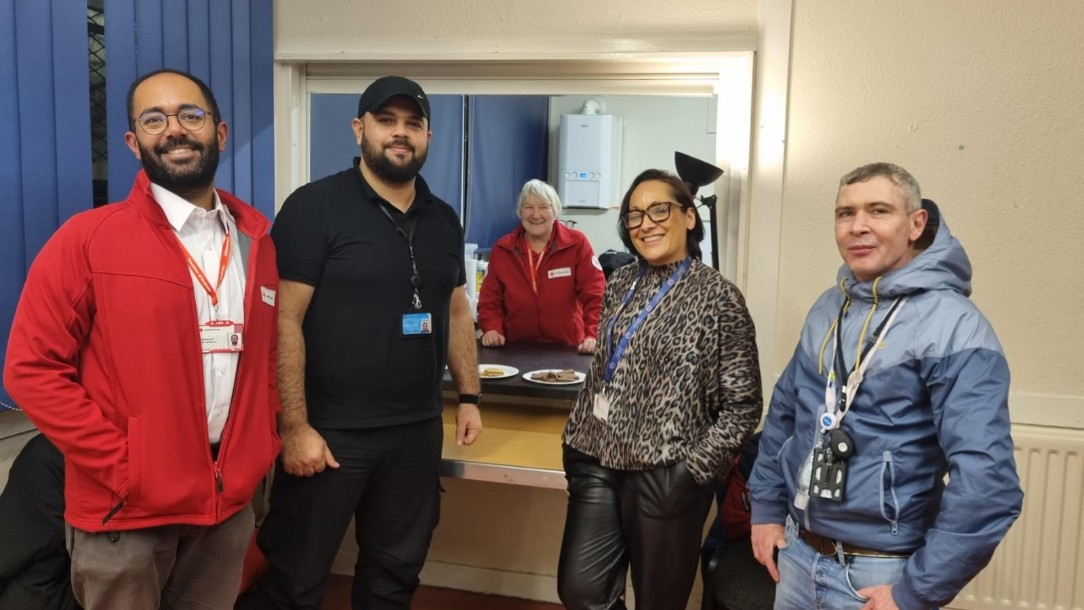 Five people, two in British Red Cross uniforms, look at the camera and smile. They are standing inside a well-lit room, which is part of a cold weather shelter in Southwark, South London