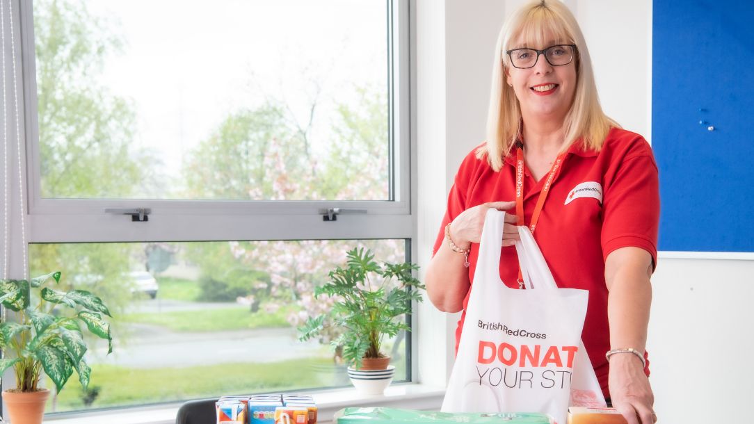 Volunteer Christine stands over a food parcel and holds a bag saying 'donate to the British Red Cross.'