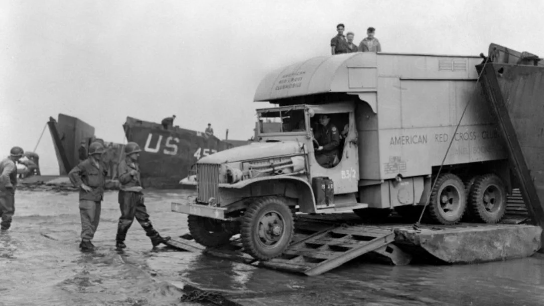 A black and white photograph showing a large Red Cross truck arriving in Normandy, with soldiers looking on.