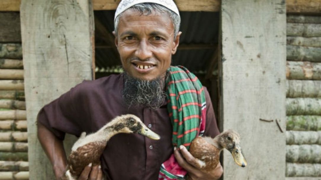 Man in Bangladesh holds two small ducks and smiles at the camera.