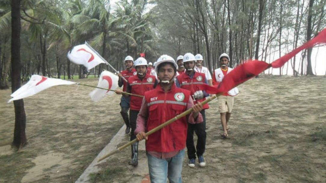 A group of Red Crescent volunteers walking through some woods in Bangladesh, waving Red Crescent flags