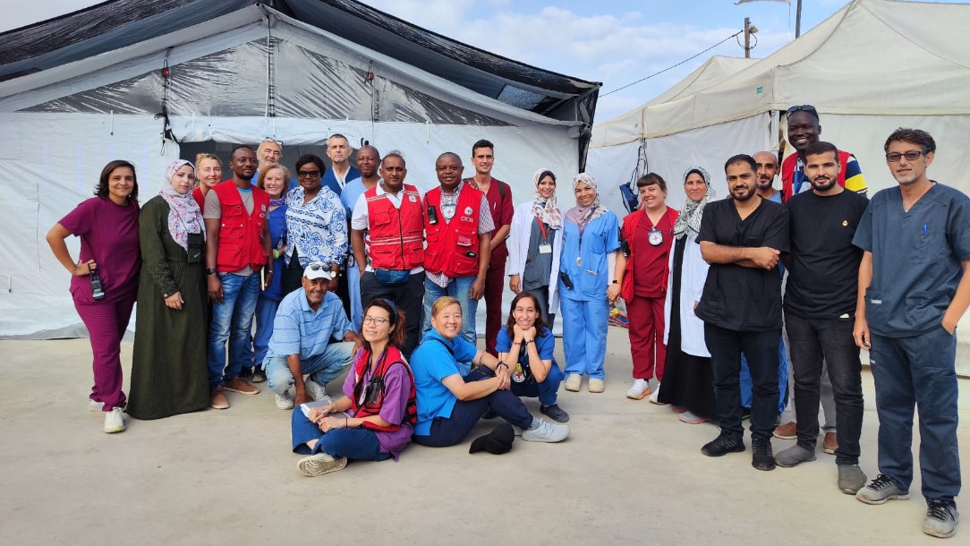 A group of medical staff stand looking to camera in front of two large tents. Some wear Red Cross uniforms and some wear surgical scrubs.