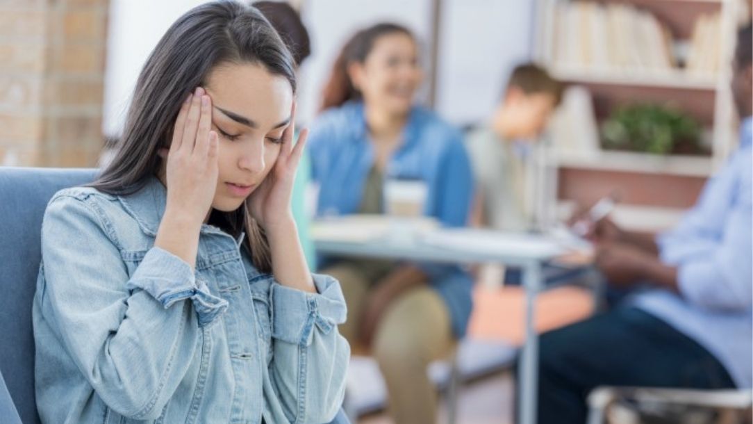 A young woman holds her head in pain while surrounded by other students.