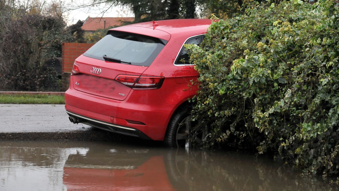 A red car seen haphazardly landed in a bush, surrounded by flood water