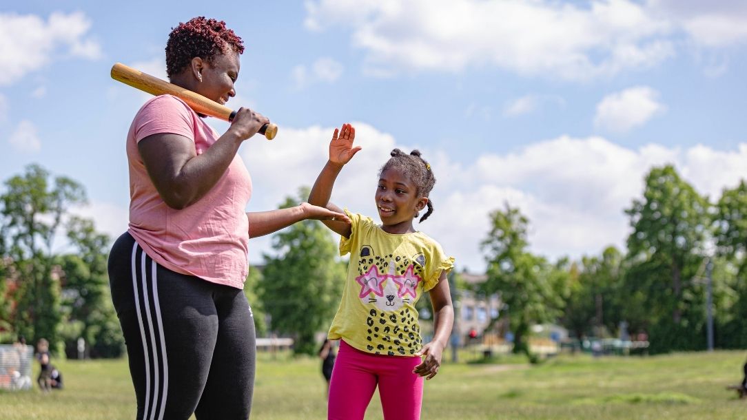 A mother and child stand in a park smiling, about to play baseball.