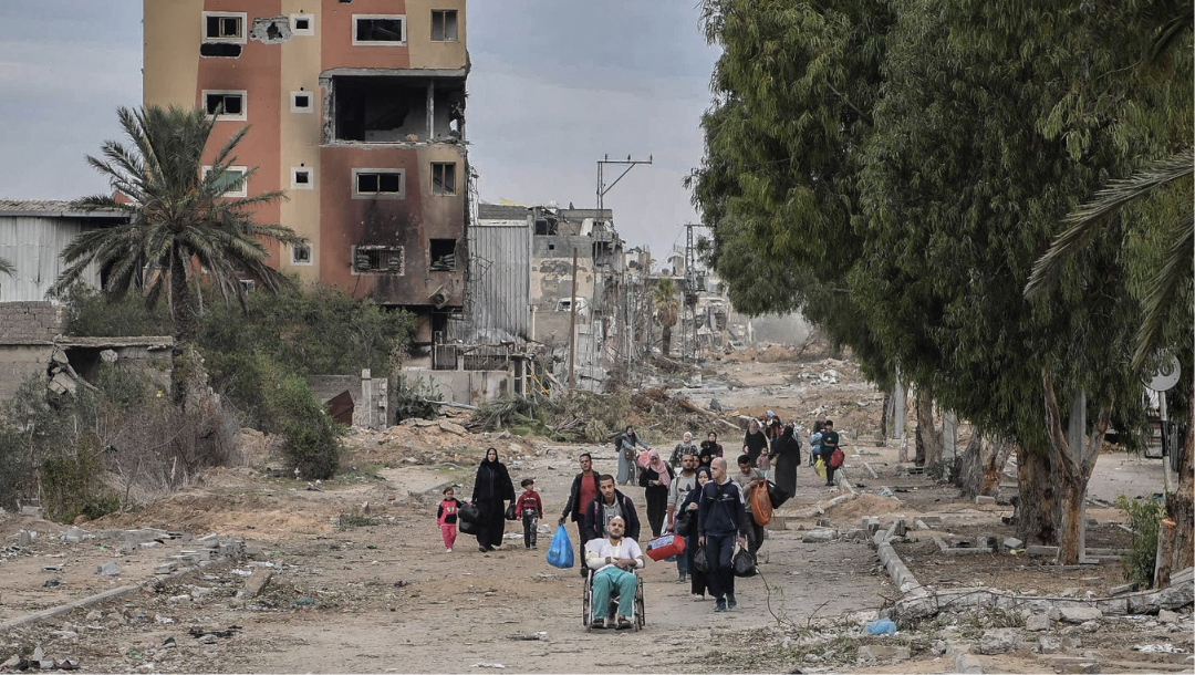 Civilians, including children and wheelchair users, holding possessions walking down a dusty path, with destroyed buildings and rubble in the background. They have been forcibly displaced from Gaza City to the southern Gaza Strip on the second day of the declared 4-day truce in Gaza City.