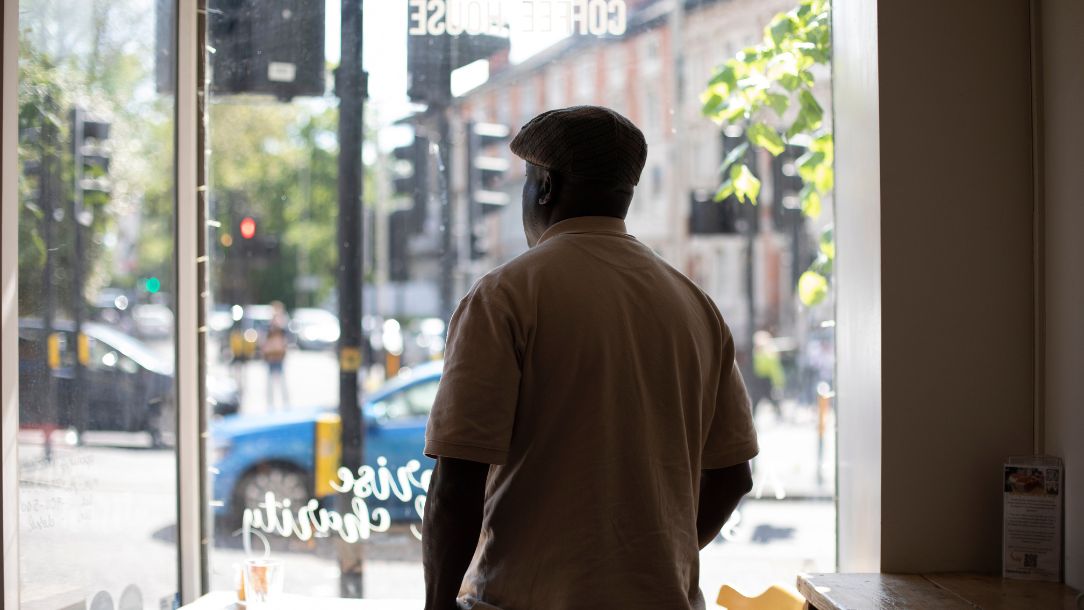George, an ambassador from the British Red Cross VOICES network, wears a flat cap stares out of a window with his back to the camera.