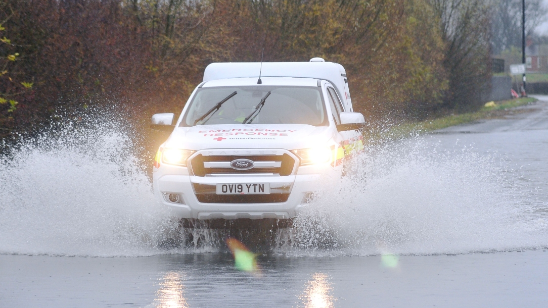 A British Red Cross emergency vehicle drives through water that has pooled on a road. 