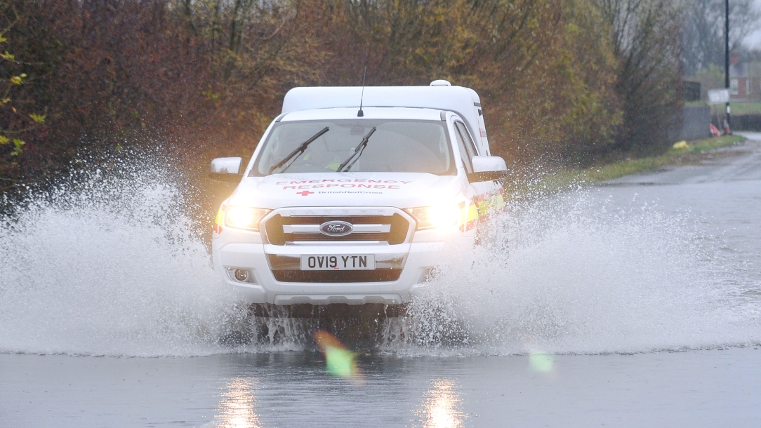A British Red Cross emergency vehicle drives through water that has pooled on a road.