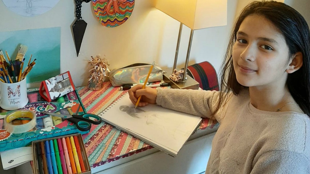 A girl sits at a table drawing with coloured pencils and looking at the camera.