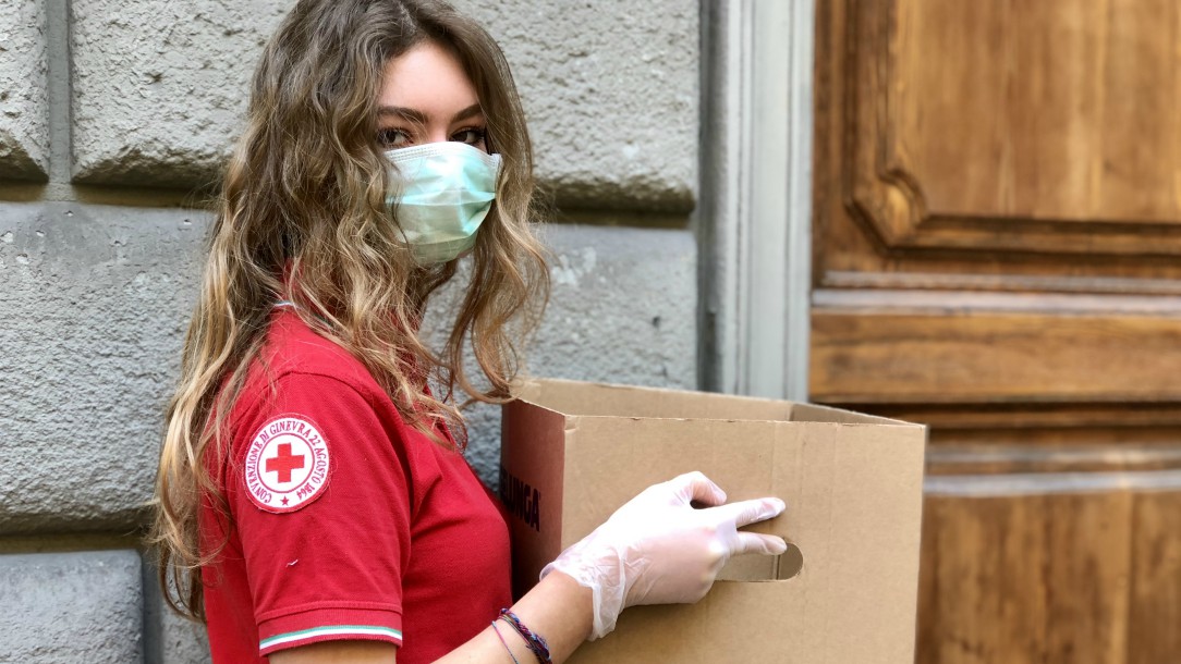 A Red Cross volunteer wears a mask and carries a cardboard box outside a door to a building.