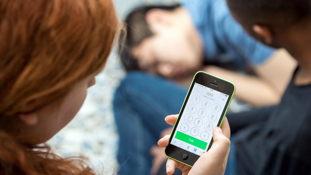 A girl calls 999 on her mobile phone for first aid advice as a young man lies on the ground with his eyes closed.