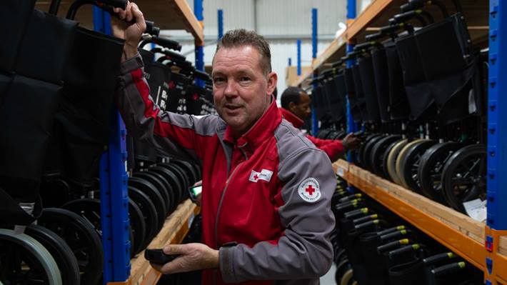 A British Red Cross volunteer stands in a warehouse with shelves of wheelchairs behind him.