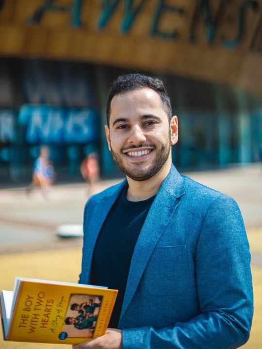 A photo of author Hamed Amiri with his book, Boy with two hearts, standing in front of Cardiff stadium.