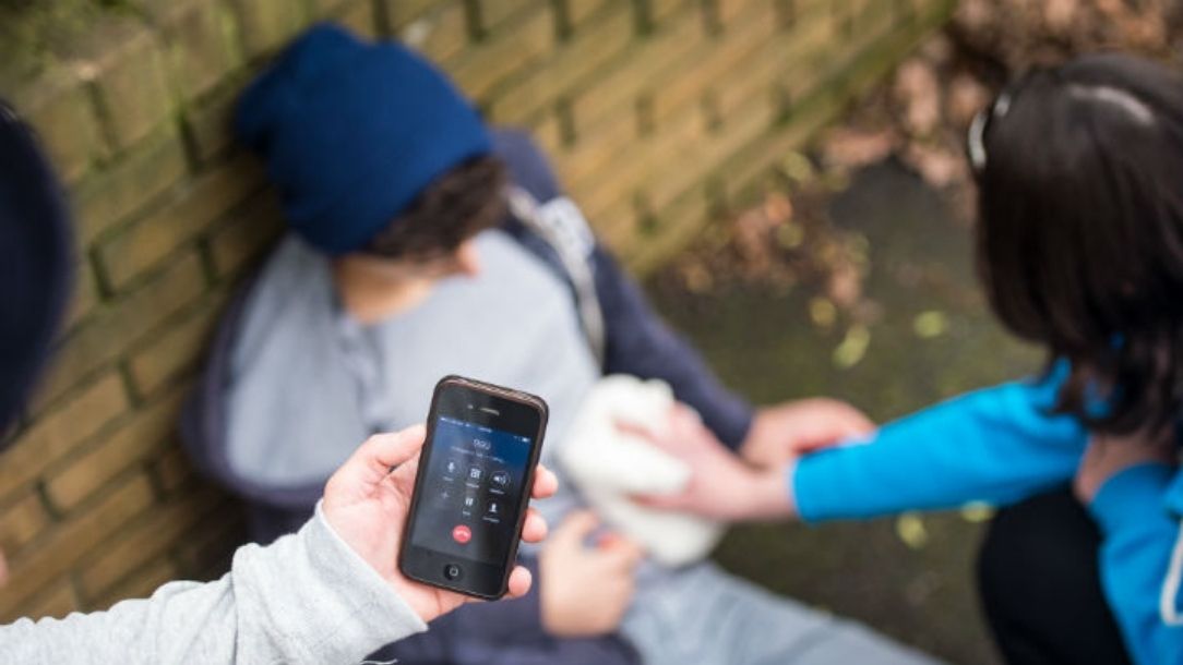 Two adults help a young man who has been stabbed, one applies pressure to the wound while the other calls emergency services.